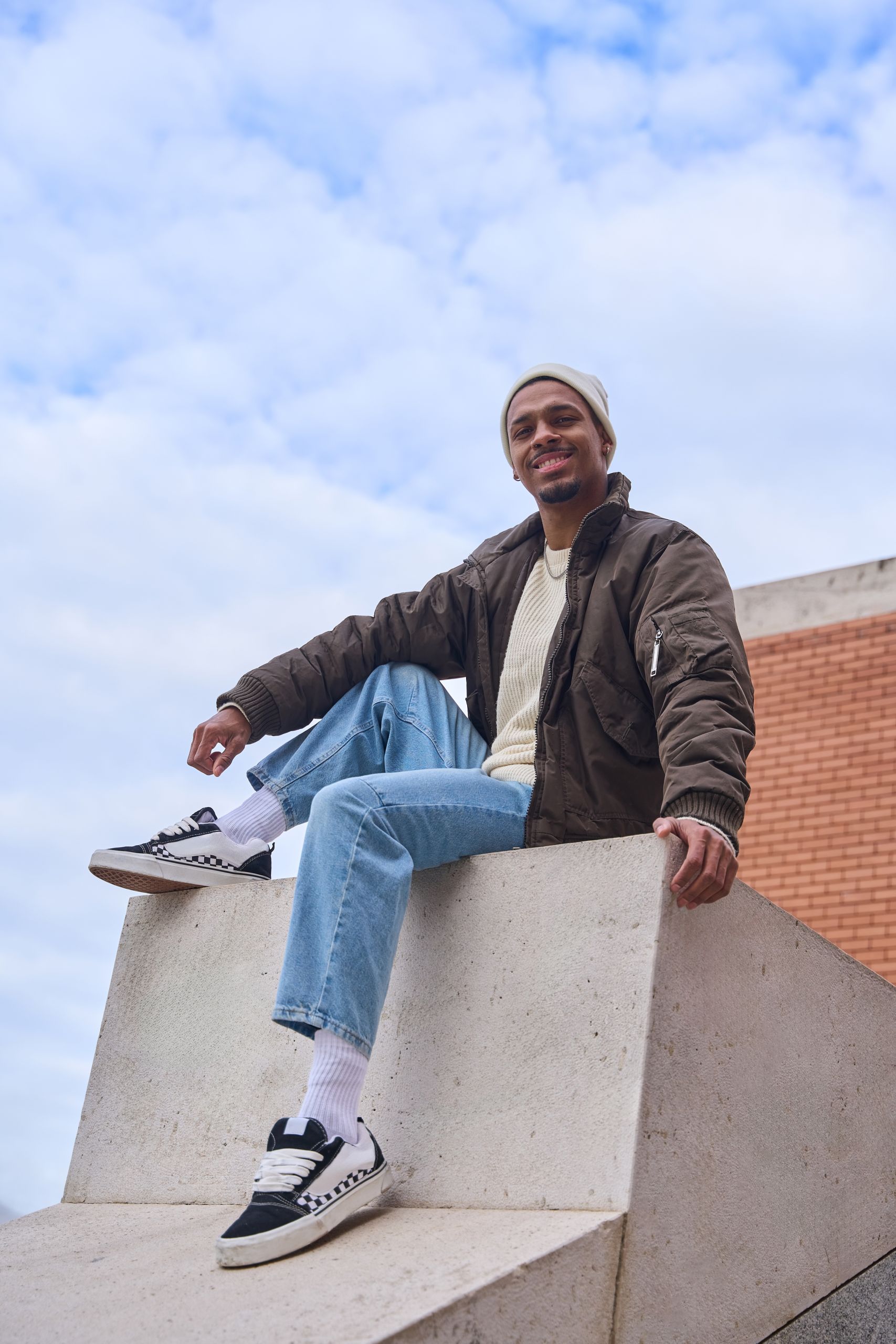 Portrait of a young man wearing a brown jacket, light blue jeans, sneakers and a white beanie, sitting on a concrete block in a modern urban setting